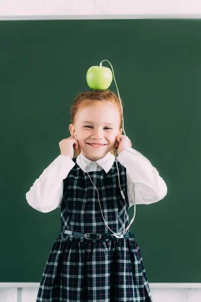 Adorable little schoolgirl with apple on head and earphones smiling and looking at camera — Stock Photo