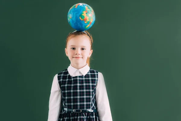 Adorable little schoolgirl with globe on head standing near blackboard and smiling at camera — Stock Photo