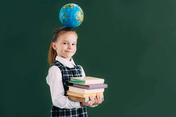 Красивая маленькая школьница с глобусом на голове держа кучу книг и улыбаясь в камеру — стоковое фото