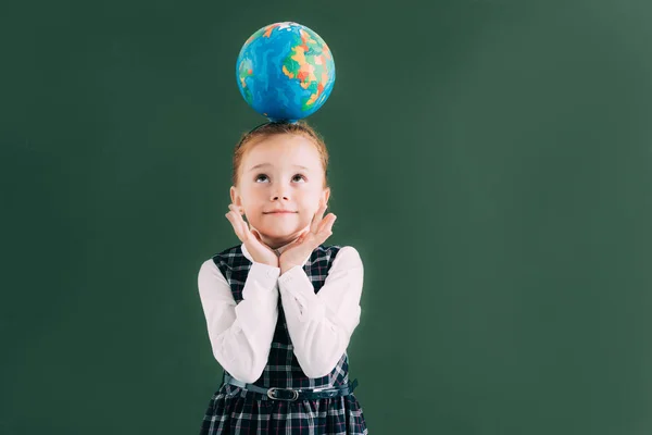 Чарівна маленька руда школярка з глобусом на голові, дивлячись вгору, стоячи біля дошки — стокове фото
