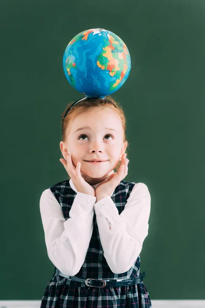 Mignonne écolière souriante avec globe sur la tête levant les yeux tout en se tenant près du tableau noir — Photo de stock