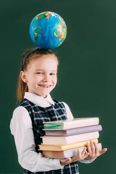 Adorable little schoolchild with globe on head holding pile of books and smiling at camera — Stock Photo