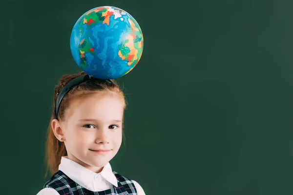 Adorable little schoolchild with globe on head smiling at camera while standing near blackboard — Stock Photo