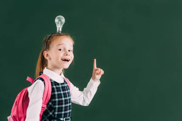 Entzückend glückliches Schulkind mit Rucksack und Glühbirne auf dem Kopf zeigt mit dem Finger nach oben und lächelt in die Kamera — Stockfoto