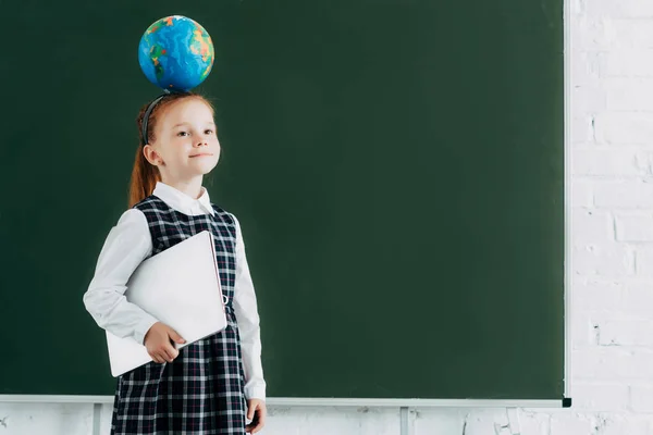 Belle petite écolière avec globe sur la tête tenant un ordinateur portable et debout près du tableau — Photo de stock