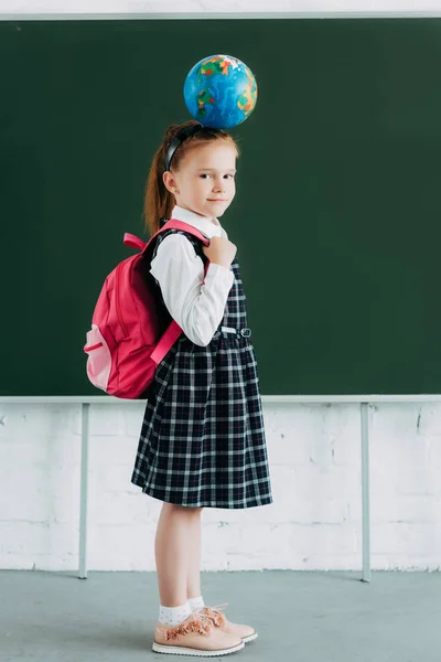 Adorável colegial com mochila e globo na cabeça sorrindo e olhando para a câmera — Fotografia de Stock