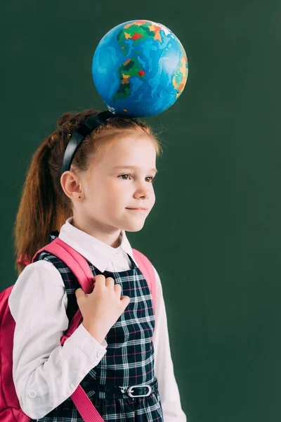 Entzückendes kleines Schulmädchen mit Rucksack und Globus auf dem Kopf, das wegschaut — Stockfoto