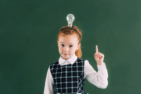 Adorabile studentessa rossa con lampadina sulla testa rivolta verso l'alto con il dito e guardando la fotocamera — Foto stock