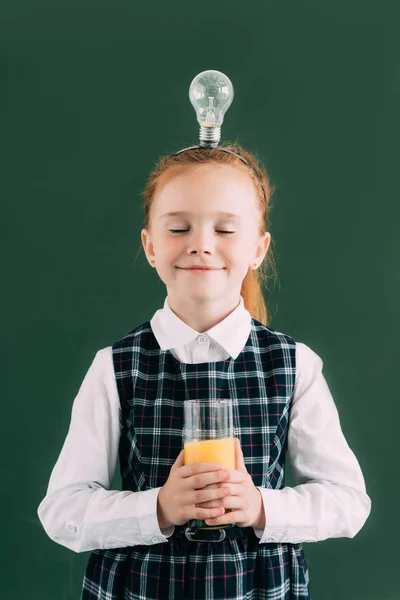 Happy little schoolgirl with closed eyes and light bulb on head holding glass of orange juice — Stock Photo