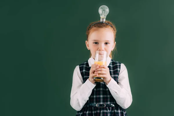 Adorable little schoolgirl with light bulb on head drinking juice and looking at camera — Stock Photo