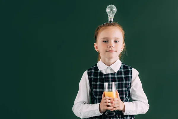 Adorable little schoolgirl with light bulb on head holding glass of juice and smiling at camera — Stock Photo