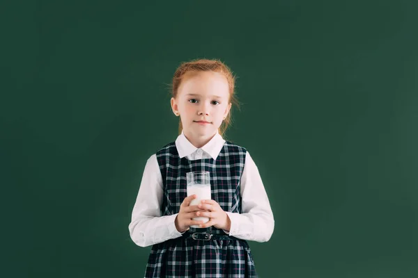 Adorable little schoolgirl holding glass of milk and smiling at camera while standing near blackboard — Stock Photo