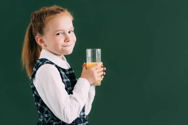 Adorable colegiala sosteniendo un vaso de jugo de naranja y sonriendo a la cámara - foto de stock