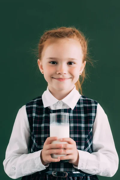Adorable colegiala sosteniendo un vaso de leche y sonriendo a la cámara - foto de stock