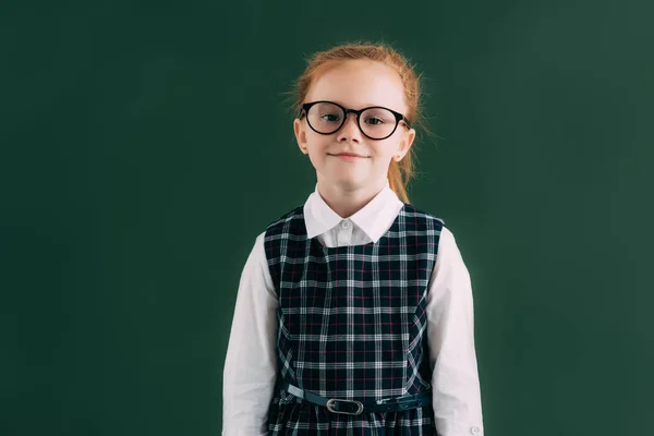 Entzückendes kleines Schulmädchen mit Brille, das neben der Tafel steht und in die Kamera lächelt — Stockfoto