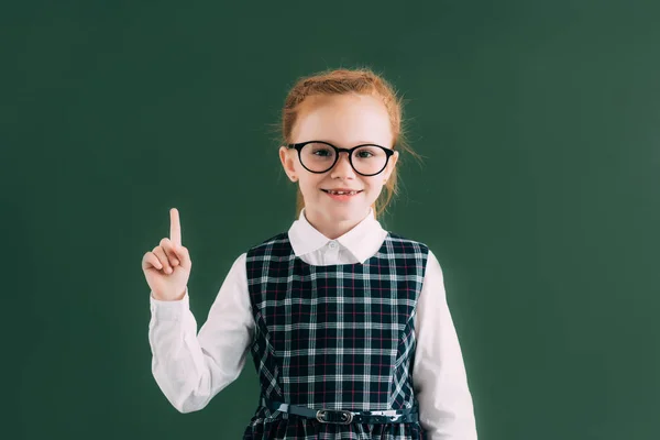 Adorable petite écolière aux lunettes pointant du doigt et souriant à la caméra tout en se tenant près du tableau noir — Photo de stock