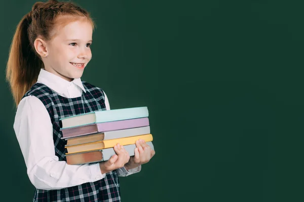 Bela sorrindo pequena estudante segurando pilha de livros e olhando para longe enquanto estava perto de quadro negro — Fotografia de Stock