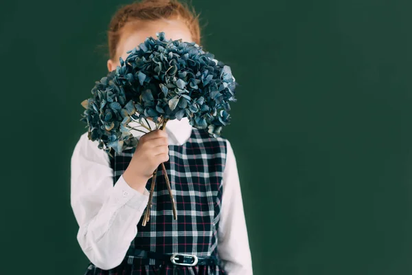 Beautiful little schoolgirl holding blue flowers on twigs while standing near chalkboard — Stock Photo