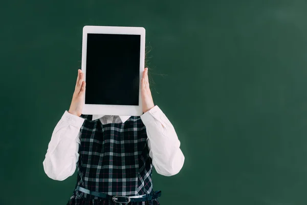 Schoolchild hiding face with digital tablet while standing near chalkboard — Stock Photo