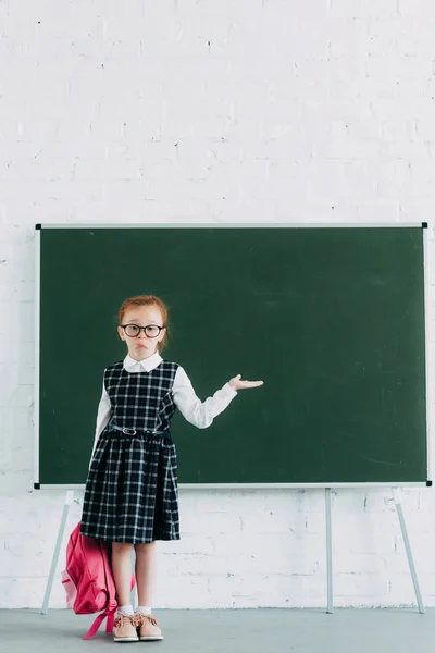 Adorale petite écolière en lunettes tenant sac d'école rose et montrant tableau blanc — Photo de stock