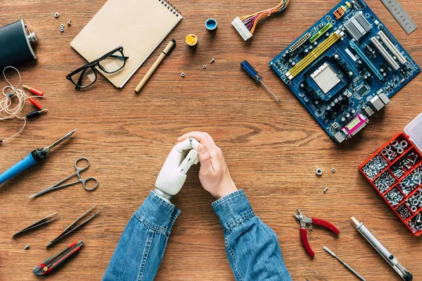 Imagen recortada de ingeniero electrónico con mano robótica sentado a la mesa con instrumentos y placa base - foto de stock