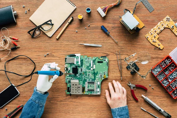 Cropped image of electronic engineer with robotic hand fixing motherboard by soldering iron at table surrounded by tools — Stock Photo