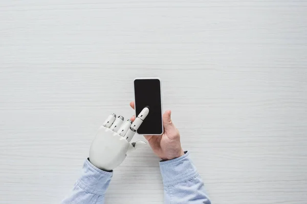 Обрезанное изображение человека с киборгом руки с помощью смартфона с пустым экраном над деревянным столом — стоковое фото