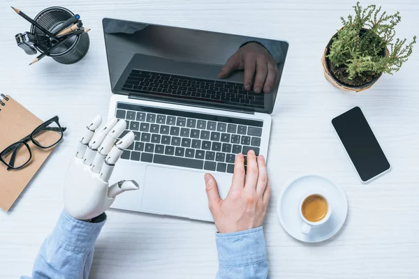 Cropped image of businessman with prosthetic arm using laptop at table with potted plant and cup of coffee — Stock Photo