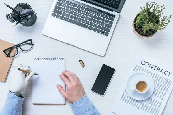 Cropped image of businessman with prosthetic arm writing in textbook at table with contract papers and gadgets in office — Stock Photo