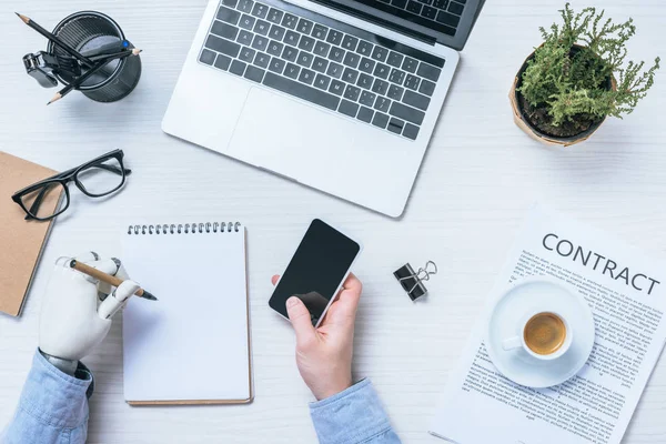 Partial view of businessman with prosthetic arm using smartphone and writing in textbook at table in office — Stock Photo