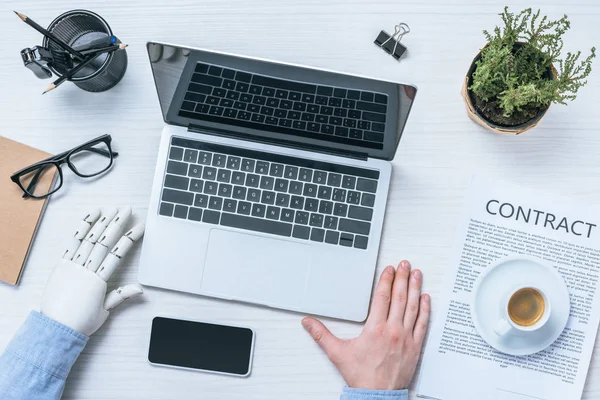 Vista parcial del empresario con brazo protésico trabajando en la mesa con smartphone, laptop y papeles de contrato en la oficina - foto de stock