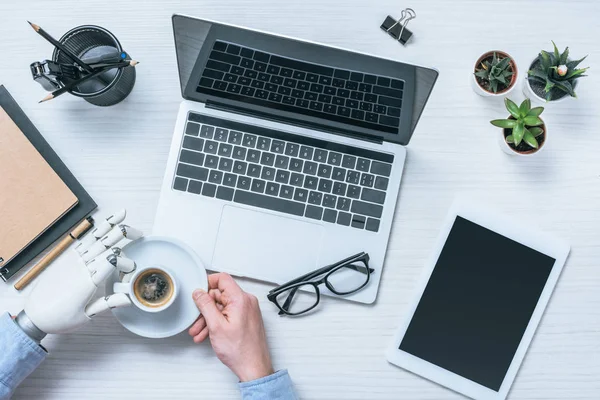 Imagem recortada de empresário com braço protético bebendo café à mesa com laptop, tablet digital e óculos — Fotografia de Stock