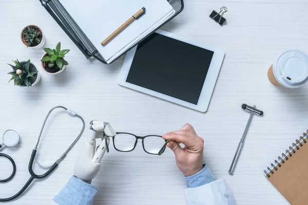 Imagen recortada del médico masculino con prótesis del brazo que sostiene las gafas en la mesa con la tableta digital en blanco — Stock Photo