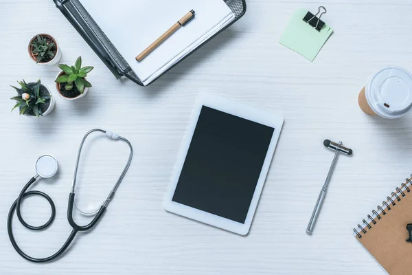 Vista superior de la tableta digital con pantalla en blanco, estetoscopio, martillo reflejo y taza de papel de café en la mesa del médico - foto de stock