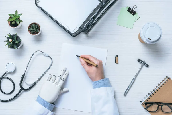 Обрізане зображення чоловіка-лікаря з протезною рукою, що пише на порожньому папері за столом з рефлекторним молотком, стетоскопом та чашкою кави — стокове фото