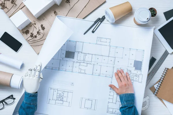 Image recadrée de l'architecte masculin avec bras prothétique travaillant avec le plan à la table avec smartphone et modèle de maison — Photo de stock