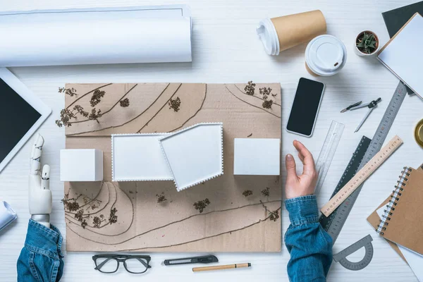 Cropped image of male architect with prosthetic arm making model of house at table with coffee cups, rulers and blueprint — Stock Photo