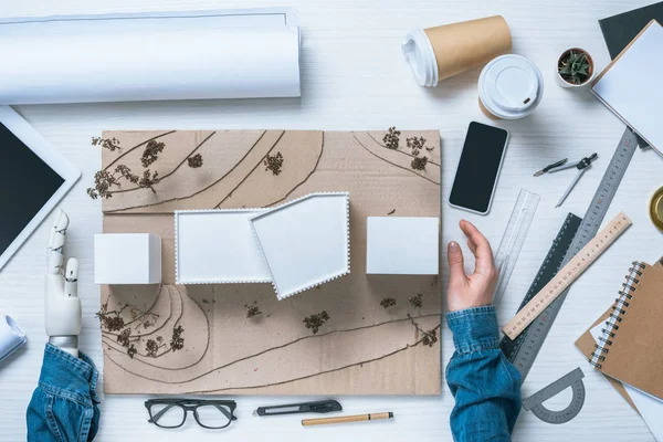 Image recadrée de l'architecte masculin avec prothèse bras faisant modèle de maison à table avec tasses à café, règles et plan — Photo de stock