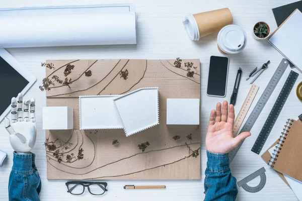 Image recadrée de l'architecte masculin avec prothèse bras faisant modèle de maison à table avec smartphone, plan et tablette numérique — Photo de stock