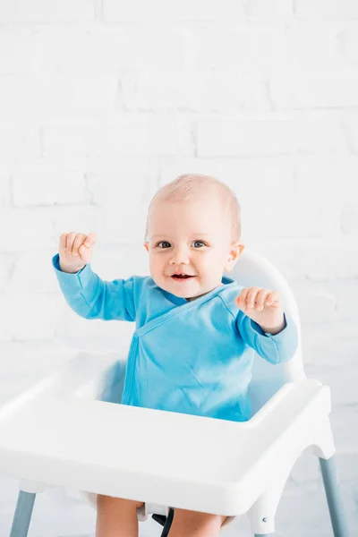 Adorabile piccolo bambino seduto sul seggiolone davanti al muro di mattoni bianchi alzato le mani — Foto stock