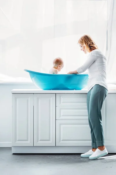 Vista lateral de la madre bañando al niño en la bañera de plástico en casa - foto de stock