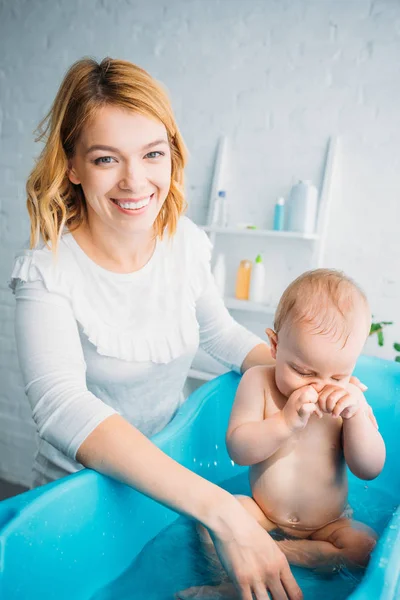 Feliz madre bañando al niño en la bañera de plástico en casa y mirando a la cámara - foto de stock