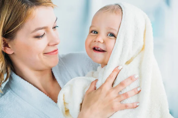 Sonriente madre en albornoz sosteniendo feliz niño pequeño cubierto con toalla - foto de stock