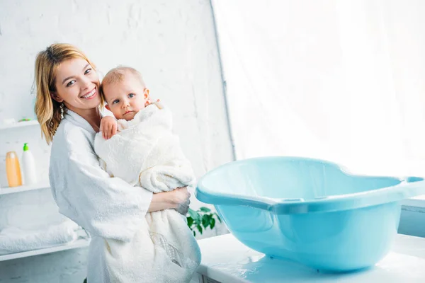 Sonriente madre en albornoz llevando adorable niño cubierto de toalla cerca de la bañera de plástico del bebé - foto de stock