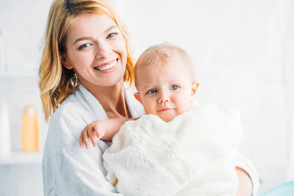 Улыбающаяся мать в халате держит маленького ребенка покрытого полотенцем и смотрит в камеру — стоковое фото