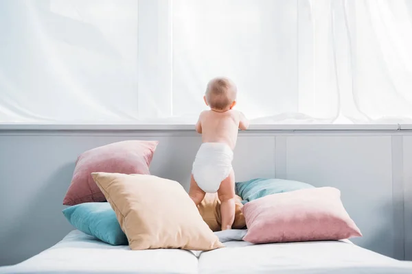 Bebê pequeno feliz em fralda de pé na cama com muitos travesseiros e olhando através da janela — Fotografia de Stock