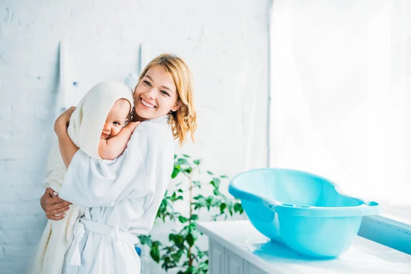 Happy mother in bathrobe carrying adorable child covered in towel near plastic baby bathtub — Stock Photo