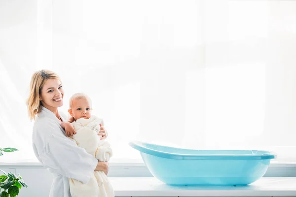 Vista laterale della madre felice in accappatoio con adorabile bambino coperto di asciugamano vicino alla vasca da bagno in plastica per bambini — Foto stock
