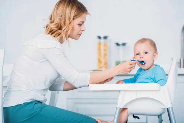 Beautiful young mother feeding child while he sitting in high chair at kitchen — Stock Photo