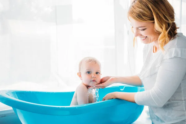 Vue latérale de mère heureuse lavant son adorable petit enfant dans une baignoire en plastique pour bébé à la maison — Photo de stock